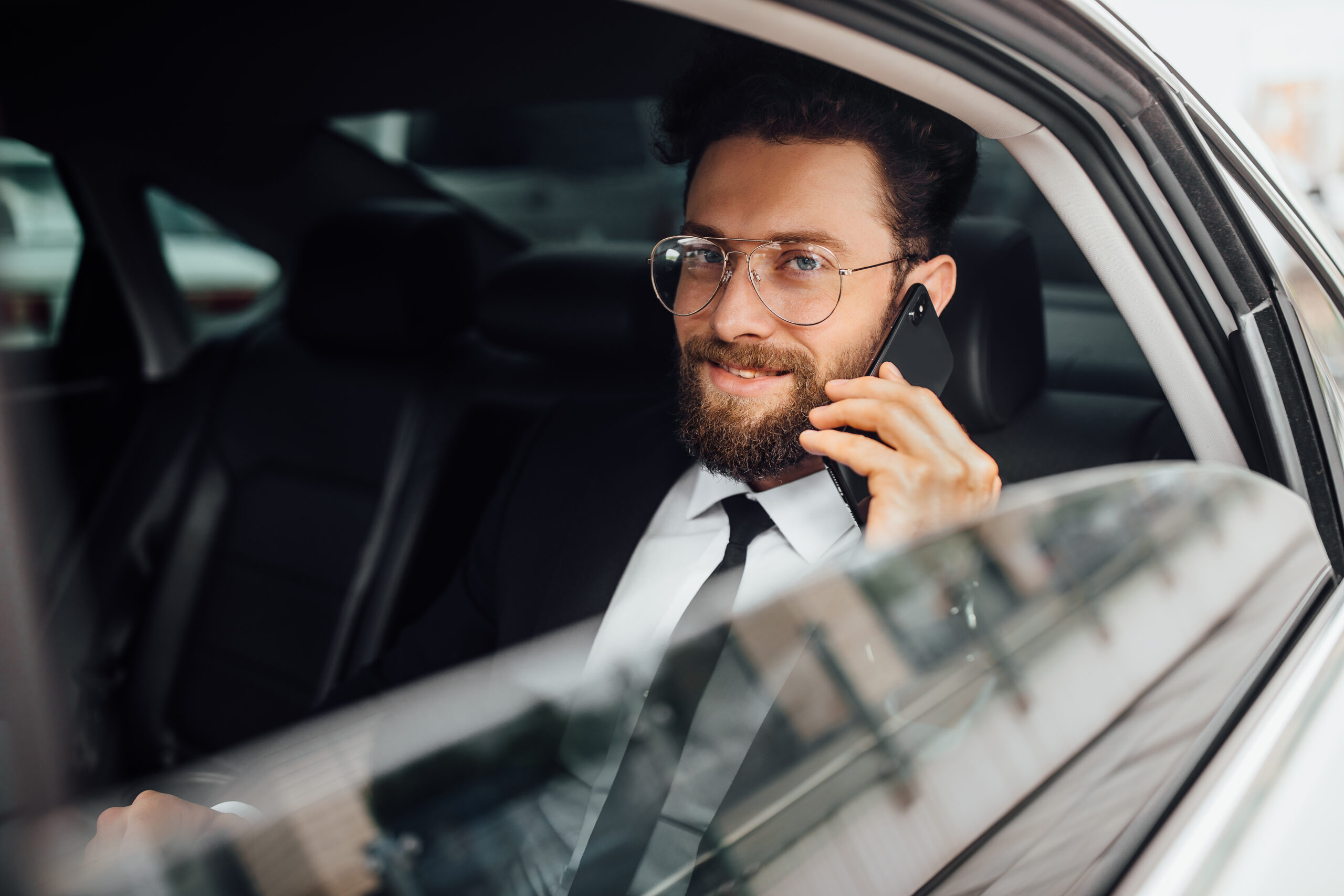 handsome-bearded-smiling-businessman-in-black-suit-calling-on-the-phone-on-backseat-of-the-car